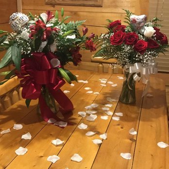 Two red rose arrangements with satin bows and scattered rose petals on a pine cabin table in Sevierville, Tennessee