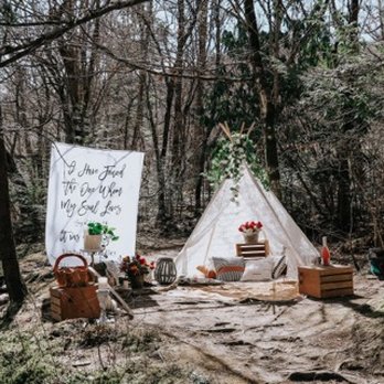 Outdoor boho picnic setup with a canvas teepee, banner, wooden crates and florals in a wooded clearing